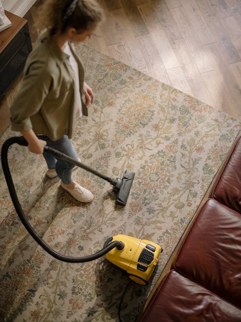 A professional cleaner dressed in white protective gear, including a full-body suit, gloves, mask, and goggles, is performing surface cleaning in a residential living room. They are using a vacuum or 