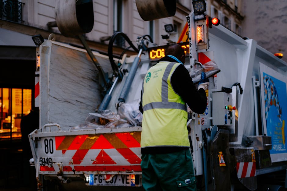 A waste collection worker wearing a high-visibility yellow vest is operating a white garbage truck on a city street at dusk. The worker, seen from behind, is handling controls on the side of the truck